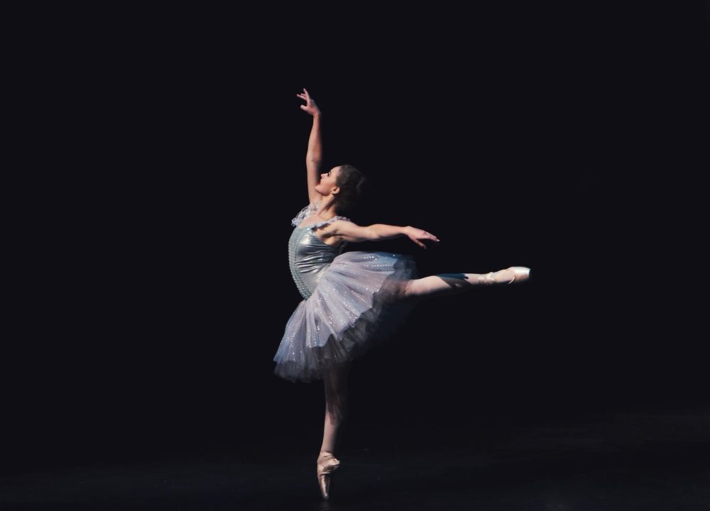 A single dancer in arabesque, very small against a large dark stage