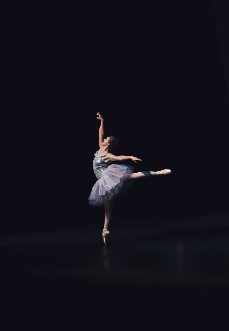 A single dancer in arabesque, very small against a large dark stage